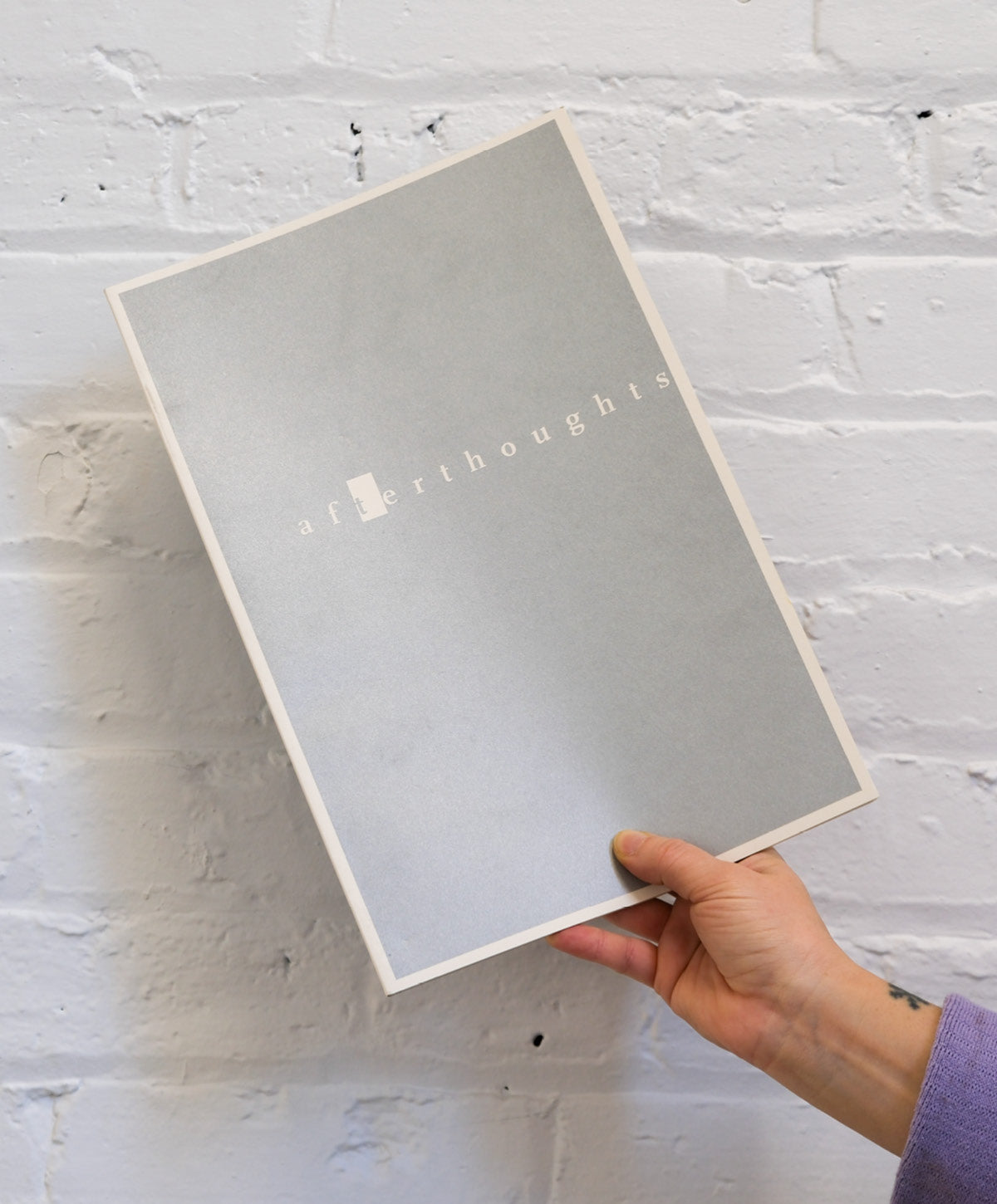 Hand holding a gray notebook with 'Thoughts' branding against a white brick wall.