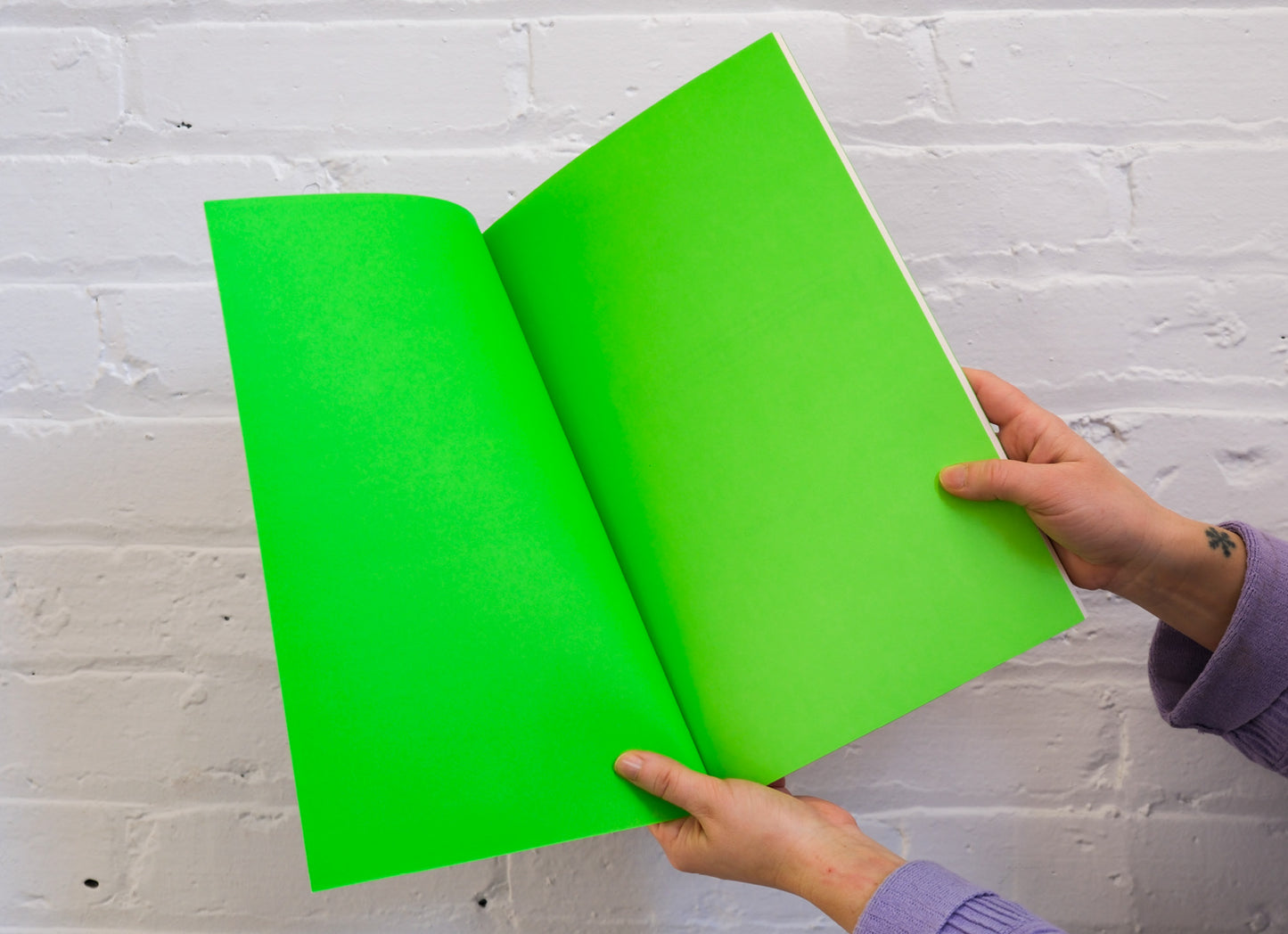 Two hands holding a bright green book spread against a white brick wall.