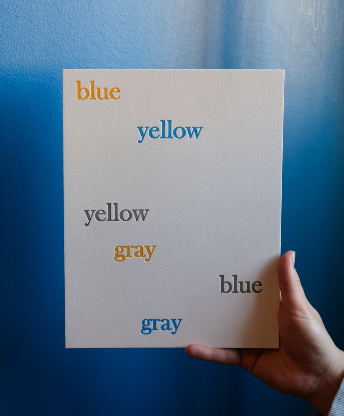 Hand holding a letterpress printed card with color swatches labelled 'blue', 'yellow', 'gray' against a blue background