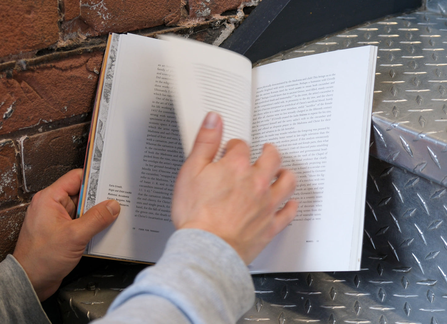 Person reading an open book on a metal surface with a brick wall background