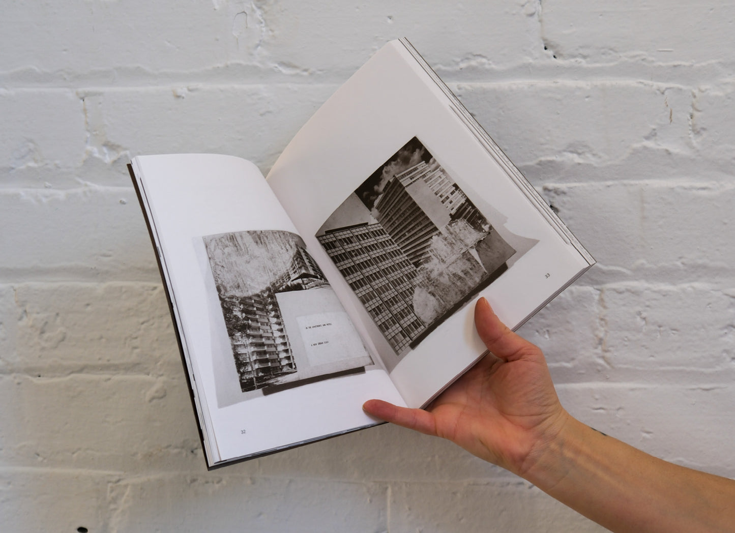 Hand holding an open book with black and white photographs against a white brick wall.