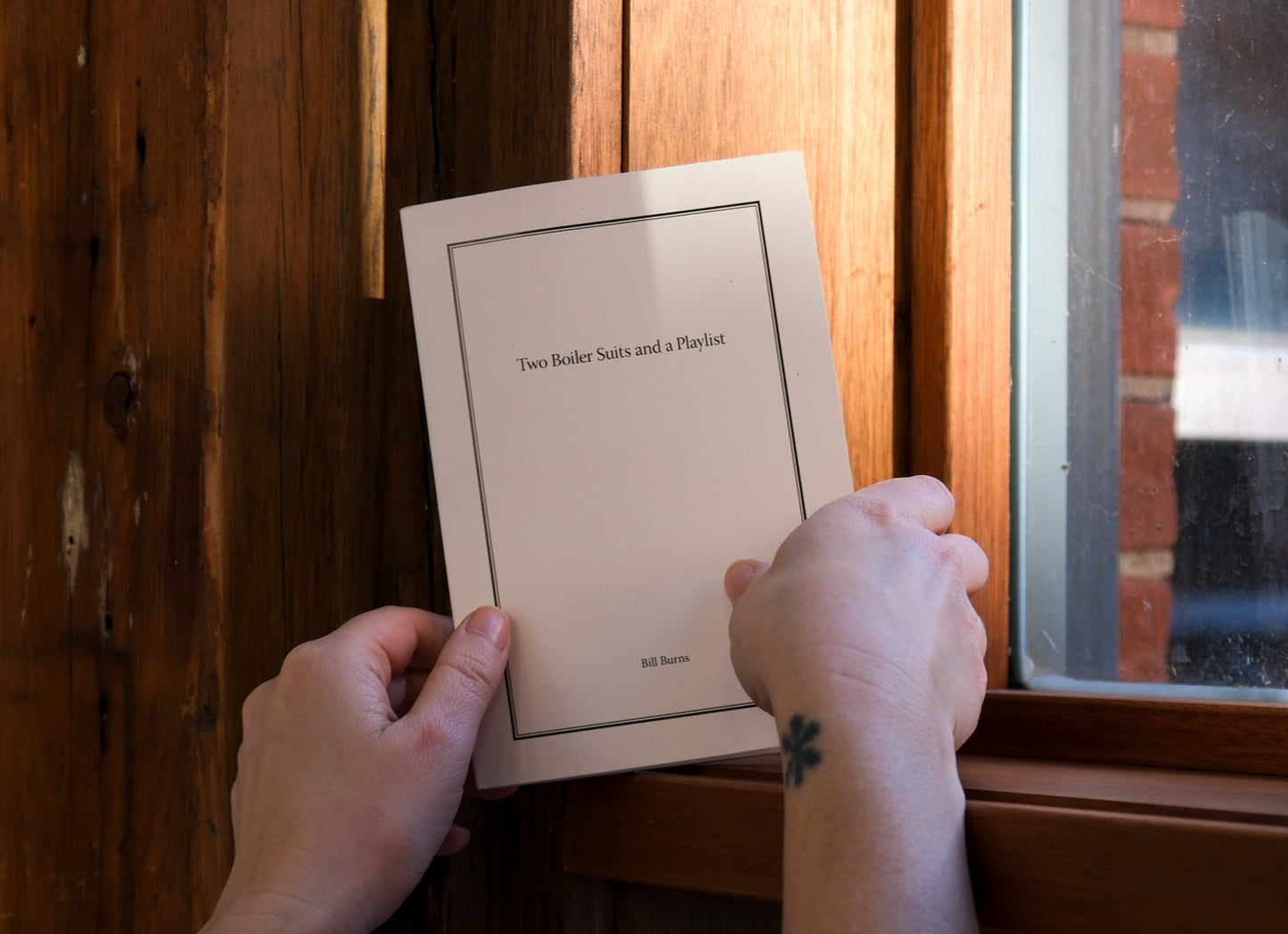 Person holding a book titled 'Two Booker Plays and a Playlist' by Willa Morris against a wooden window frame.