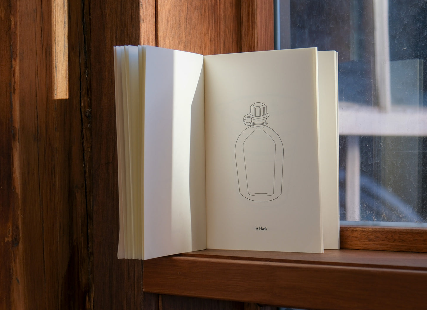 Open book with a line drawing of a bottle on a wooden surface near a window.